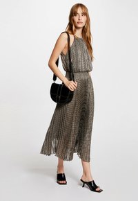 Falten-maxikleid aus schwarzem und goldenem Stoff mit hohem Ausschnitt, kombiniert mit einer schwarzen Schultertasche und offenen schwarzen Absatzmules.