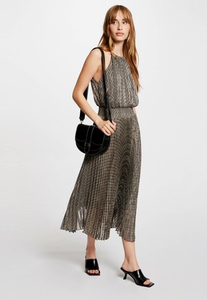 Gestreepte maxijurk van zwart en gouden stof met een hoge halslijn, gecombineerd met een zwarte schoudertas en open teen zwarte haksandalen.
