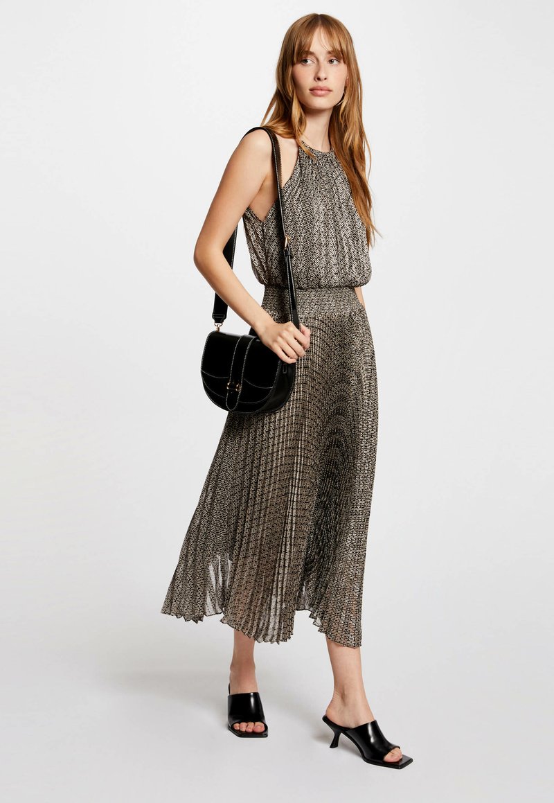 Falten-maxikleid aus schwarzem und goldenem Stoff mit hohem Ausschnitt, kombiniert mit einer schwarzen Schultertasche und offenen schwarzen Absatzmules.