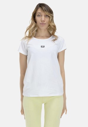 Femme aux cheveux blonds ondulés portant un t-shirt blanc avec un petit logo rectangulaire et un pantalon jaune pâle, debout devant un fond uni.