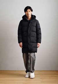 Schwarze Daunenjacke mit Kapuze, mit einem gesteppten Design und vorderen Knöpfen. Kombiniert mit grauen Jeans und weißen Sneakern auf einem Holzboden.
