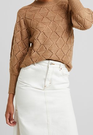 Femme portant un pull en maille marron à motif losange et une jupe en jean blanche taille haute avec une fente à l'avant.