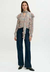 Donna in una blusa a rouches floreale pastello con un fiocco al collo, jeans neri a gamba larga e stivaletti neri a punta, in piedi di fronte a uno sfondo bianco.