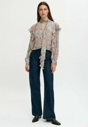 Donna in una blusa a rouches floreale pastello con un fiocco al collo, jeans neri a gamba larga e stivaletti neri a punta, in piedi di fronte a uno sfondo bianco.