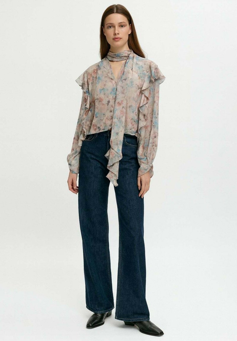 Donna in una blusa a rouches floreale pastello con un fiocco al collo, jeans neri a gamba larga e stivaletti neri a punta, in piedi di fronte a uno sfondo bianco.