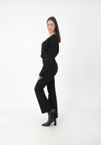 Donna in un maglione nero, pantaloni neri a vita alta e stivali neri con tacco posa di lato su uno sfondo bianco.