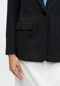 Czarny żakiet w paski z klapami, z pojedynczym zapięciem na guzik i kieszeniami z przodu. Teksturowana tkanina, dopasowany krój i długie rękawy.