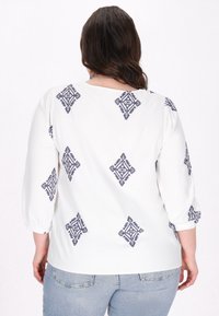 Fehér blúz sötétkék geometrikus hímzéssel, háromnegyedes ujjakkal és kerek nyakkal. Sima textúra, laza fazon.