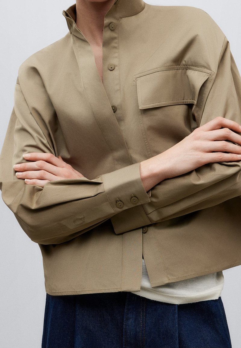 Personne portant une veste beige boutonnée avec une poche poitrine, les bras croisés sur une chemise blanche et un pantalon bleu foncé.