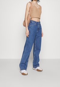Hellbraunes, kurzes Oberteil, blaue, hochgeschnittene, weit geschnittene Jeans mit kontrastierenden Nähten und weiße Sneakers mit braunen Sohlen. Schlichte Handtasche.