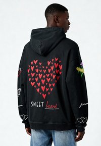 Mustmust eemel, millel on suur punane südame graafika, tekst "SWEET heart" ja lillemotiivid. Puuvillane materjal, millel on vabalt langev lõige ja soonikkoes mansetid.