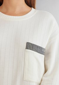 Hvid ribbet sweater med rund halsudskæring og en lomme, der har et sort houndstooth-mønster som accent langs den øverste kant. Blødt, struktureret stof.
