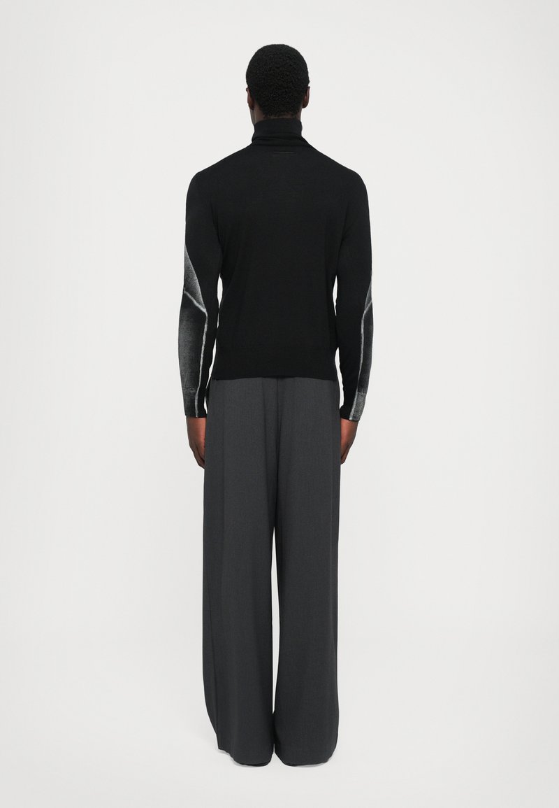Uomo vestito con un dolcevita nero con design astratti grigi sulle maniche e pantaloni larghi grigio scuro, in piedi rivolto di spalle.