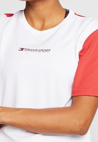 Osoba oblečená v bílém a červeném sportovním tričku s krátkým rukávem a logem "Tommy Sport" na hrudi.