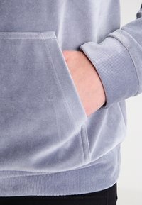 Šedá sametová mikina s kapucí, která má přední kapsu, žebrované manžety a jemnou texturu. Detailní pohled zaměřený na kapsu a oblast zápěstí.