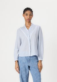 Camicia a righe blu e bianche con colletto, fronte plissettato e maniche lunghe, abbinata a pantaloni chiari in denim testurizzati.