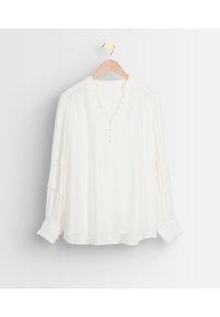 Vit långärmad blus med framknappar, plisserade detaljer på ärmarna och manschetterna, hängande på en trägalje mot en vit bakgrund.