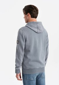 Felpa grigia con cappuccio in misto cotone, con polsini e orlo a costine, e un dettaglio patch sulla manica sinistra. Indossata con jeans blu.