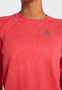 Červená košile s dlouhým rukávem vyrobená z texturované tkaniny. Obsahuje žebrované detaily na ramenou a malé šedé logo na hrudi.