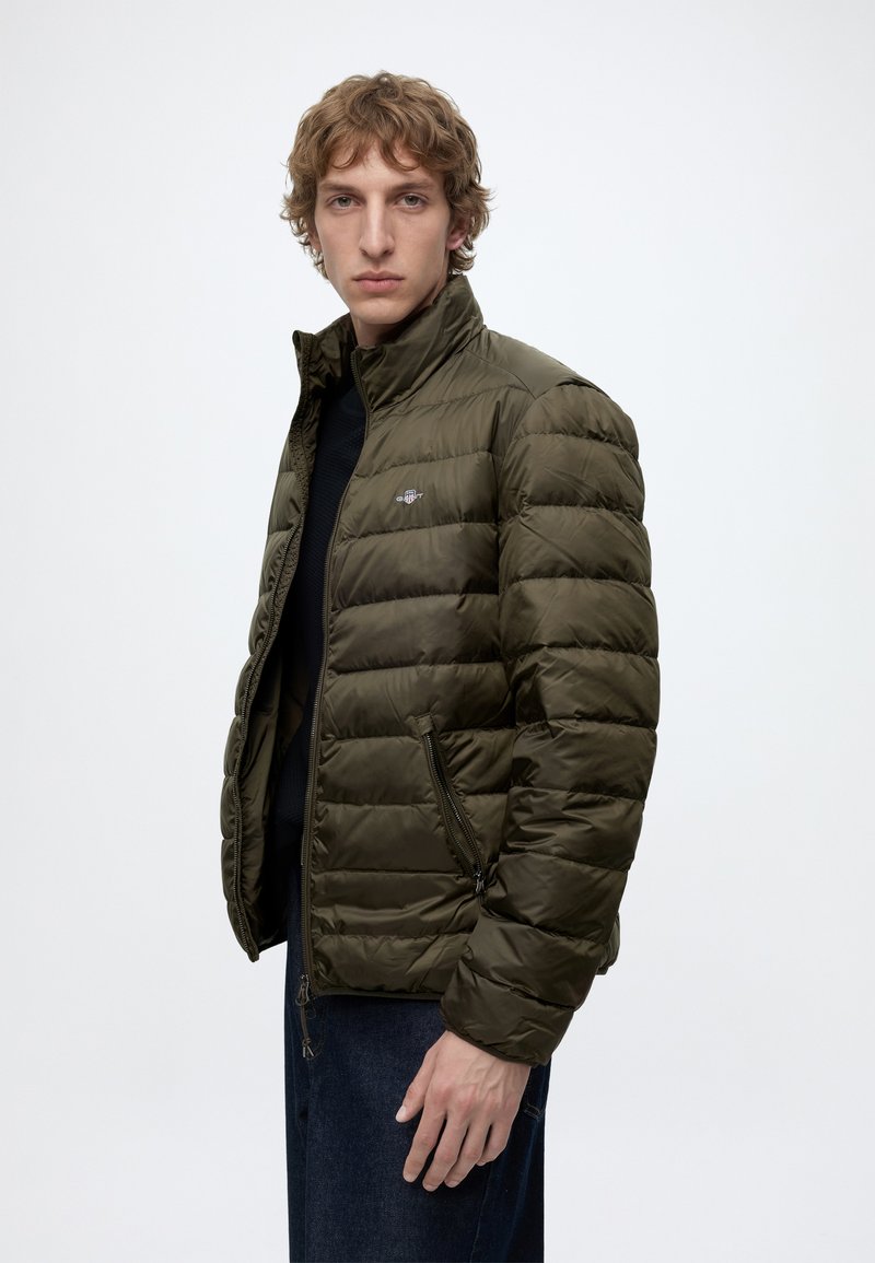 Junger Mann mit hellen lockigen Haaren, der eine olivgrüne Steppjacke und dunkle Hosen vor einem schlichten weißen Hintergrund trägt.