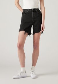 High-waisted black denim shorts with frayed hems, five-pocket design, and metal hardware accents, paired with white sneakers and socks.