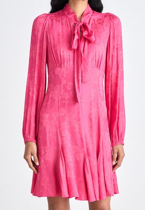 Mujer con un vestido rosa de manga larga con un sutil estampado floral, escote con lazo y pliegues ligeros en la falda.