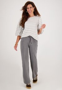 Glunderende vrouw met lang haar, die gouden ring oorbellen draagt, een wit gestreept top met pofmouwen, grijze wijde broek en bruine sneakers, die vooruit loopt.