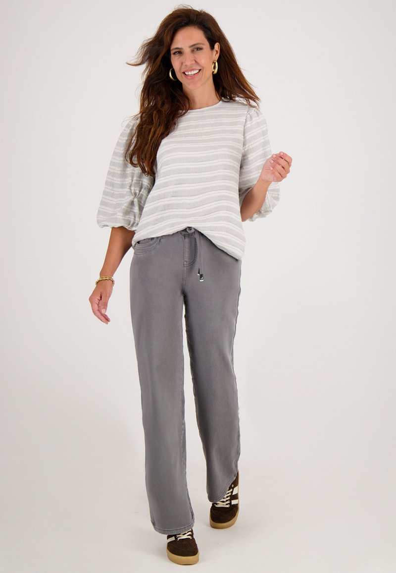 Glunderende vrouw met lang haar, die gouden ring oorbellen draagt, een wit gestreept top met pofmouwen, grijze wijde broek en bruine sneakers, die vooruit loopt.