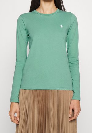 Vrouw draagt een lichtgroen shirt met lange mouwen met een klein wit logo op de borst en een geplooide lichtbruine rok.