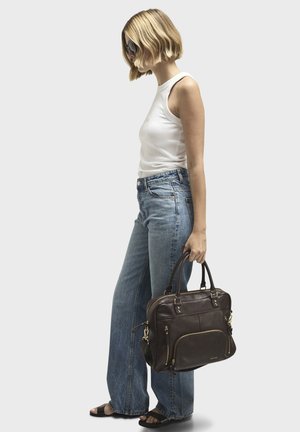 Braune Lederhandtasche mit mehreren Reißverschlüssen und Taschen, weicher Textur, robuste Griffe. Modell trägt ein weißes Tanktop und blaue Jeans.