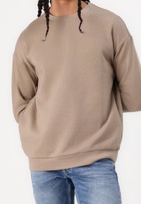 Sudadera oversize en topo con escote redondo y hombros caídos, hecha de una tela suave y texturizada. Combinada con unos vaqueros azules.