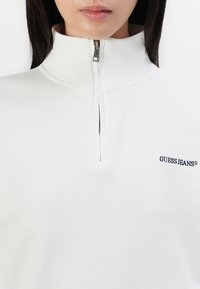 Felpa bianca con zip, caratterizzata da un colletto alto, tessuto testurizzato e un piccolo logo blu navy "GUESS JEANS" sulla parte sinistra del petto.