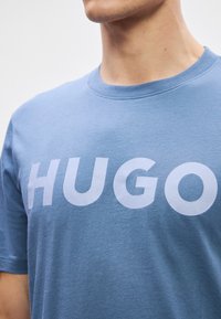 Ανοιχτό γαλάζιο βαμβακερό μπλουζάκι με κοντά μανίκια και στρογγυλή λαιμόκοψη. Διαθέτει μεγάλη λευκή εκτύπωση της λέξης "HUGO" στο στήθος.