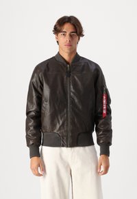 Casaco de aviador em pele castanha com punhos e bainha canelados, apresentando um fecho de correr na frente e uma etiqueta vermelha no bolso do braço esquerdo.