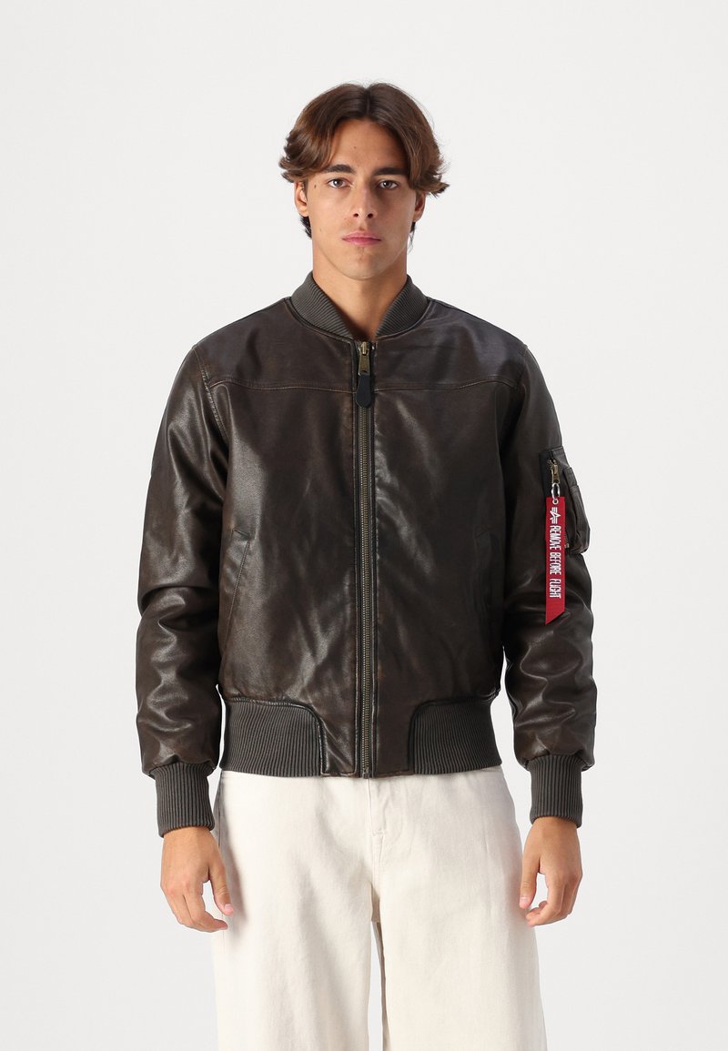 Casaco de aviador em pele castanha com punhos e bainha canelados, apresentando um fecho de correr na frente e uma etiqueta vermelha no bolso do braço esquerdo.