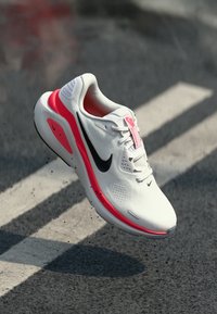 Baskets blanches avec un swoosh noir, accents roses, une tige en mesh et une semelle texturée. Design léger adapté aux sports.