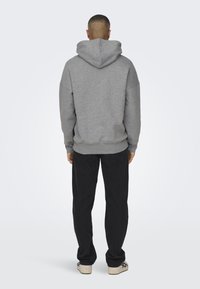 Grauer Kapuzenpullover mit großer, weiter Kapuze, lockerem Schnitt und geripptem Saum. Getragen mit schwarzen Hosen und hellen Turnschuhen.