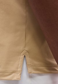 Gros plan sur un tissu beige avec un petit détail de couture fendue latérale sur un vêtement, à côté d’un bras à la peau foncée et un vêtement blanc en dessous.