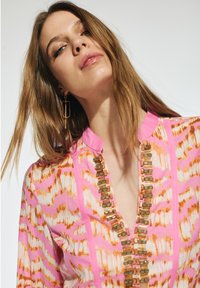 Blusa con motivo rosa e bianco con dettagli arancioni, caratterizzata da un collo a V e bottoni decorativi lungo il centro. La modella ha i capelli lunghi.