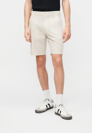 Hombre con pantalones cortos beige hasta la rodilla, calcetines blancos hasta el tobillo y zapatillas blancas con rayas negras, de pie contra un fondo liso.