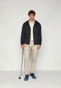 Chaqueta negra con cremallera sobre una camisa con cuello y ribetes rojos, pantalones beige y zapatillas deportivas azules; sosteniendo un palo de golf en una postura neutral.