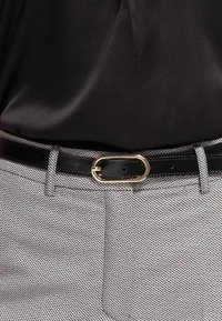 Ceinture en cuir noir avec boucle ovale dorée portée par-dessus un pantalon gris à motif chevron et une chemise en satin noir rentrée.