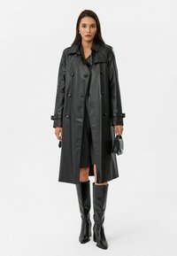 Zwarte leren trenchcoat met dubbele knopen, een kraag en zijzakken. Gekleed met zwarte kniehoge laarzen en een kleine handtas.