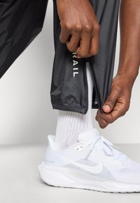 Μαύρο αθλητικό παντελόνι με φερμουάρ στο τελείωμα, συνδυασμένο με λευκά sneakers από δίχτυ και λευκές κάλτσες. Το κείμενο "TRAIL" είναι ορατό στο παντελόνι.