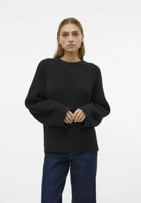 Pull en tricot noir avec une texture côtelée, des manches raglan et des poignets larges, associé à un jean bleu foncé dans un cadre minimaliste.