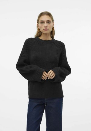 Maglione nero a maglia con una texture a coste, maniche raglan e polsini larghi, abbinato a jeans blu scuro in un ambiente minimalista.