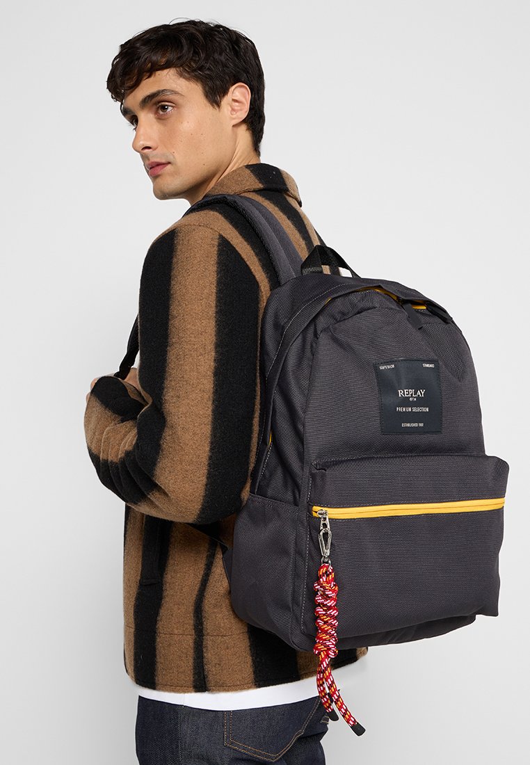 Zaino in tessuto nero con accenti gialli sulla cerniera, una tasca frontale e un portachiavi intrecciato rosso. Progettato con spallacci imbottiti.