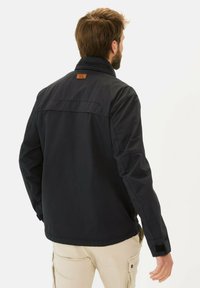 Schwarze Jacke mit hohem Kragen, aus glattem Material gefertigt. Verfügt über einen Lederaufnäher auf dem Rücken und verstellbare Klettverschlüsse an den Ärmeln.
