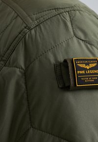 Giacca isolante verde oliva con design trapuntato, caratterizzata da un'etichetta PME Legend gialla e nera di grande rilievo, tessuto testurizzato e dettagli cuciti.