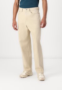 Light beige denim pants with a straight-leg fit, five-pocket design, and subtle stitching details. Worn with a blue shirt and beige sneakers.